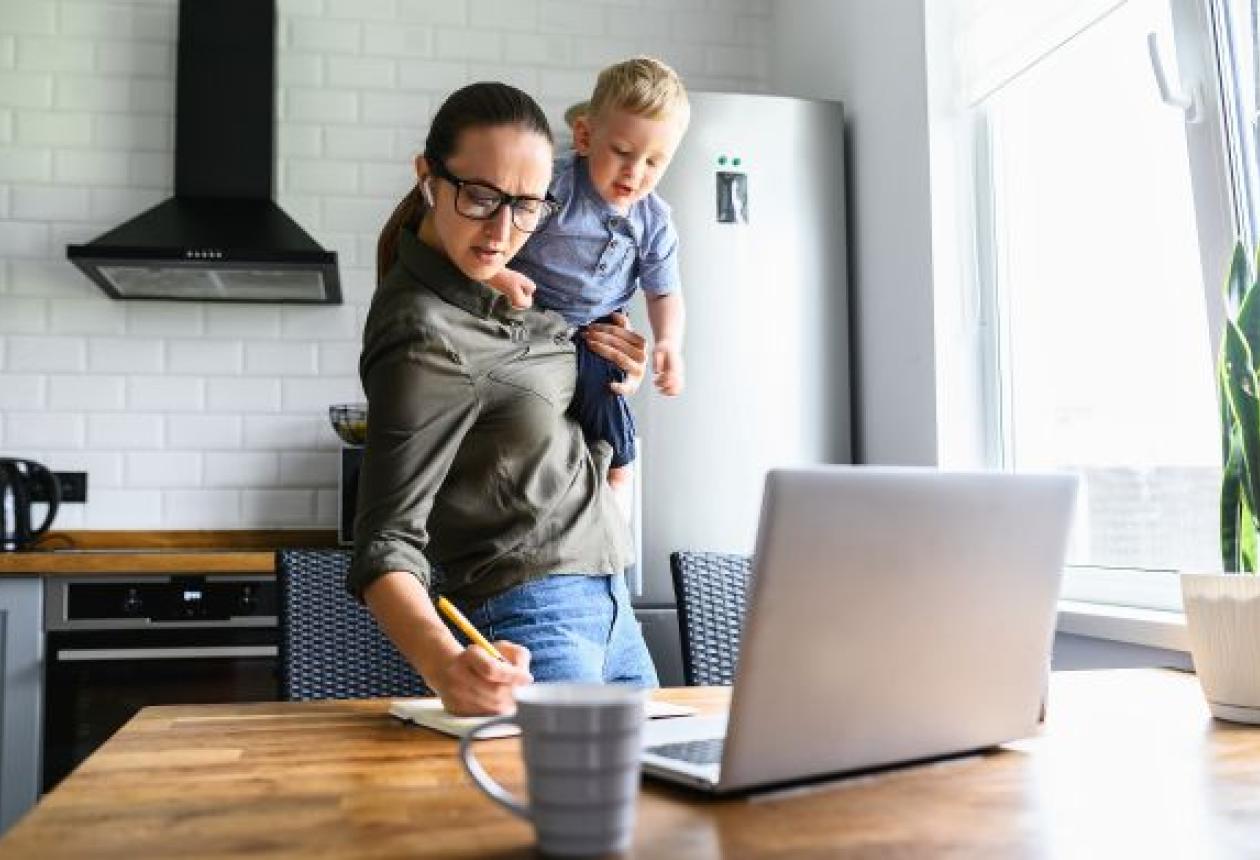 Maman débordée travaille à domicile avec son enfant actif dans les bras, prenant des notes tout en regardant son ordinateur portable.